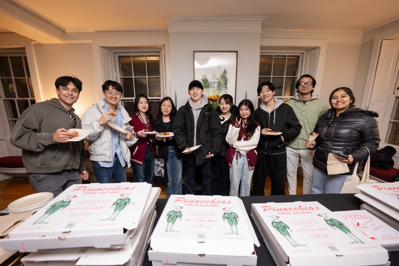 A group of students grabbing pizza at Dean Dunne's residence the night before Hosuing Day pose with their slices of Pinnochios pizza behind a row of pizza boxes. 