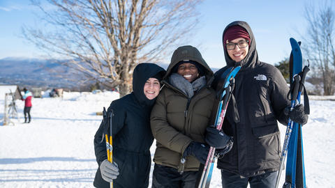 Three students pose in winter gear with skis in hand on a snowy Vermont day.