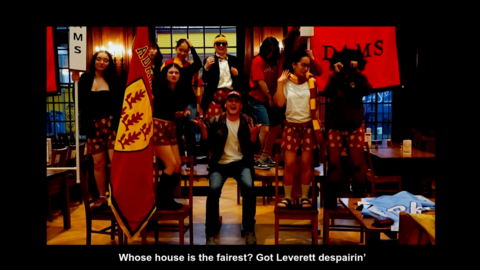 A snapshot of the Adams House Housing Day video with students holding the Adams House flag in the Adams House Dining Hall.