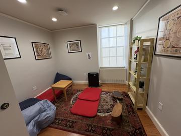The meditation space that has a rug with mats on the ground, a coffee table, a bookshelf with plants, and art on the wall.