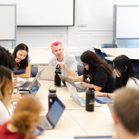 Professor at the head of the table addresses students in the classroom while most students look down at their notes.