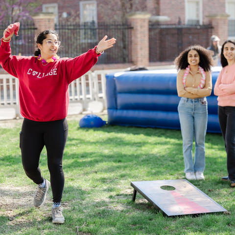 Student in a crimson sweatshirt with a Harvard College shield on it tosses a beanbag toward a corn hole board.