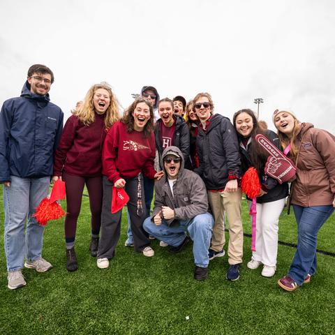 Students posing for a group photo while shouting in school spirit at the camera during the Harvard-Yale tailgate.