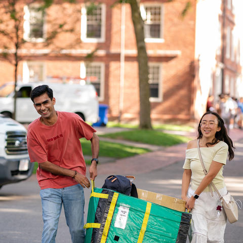 Two students carrying a large moving bin on campus. 