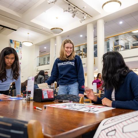 Three students at Pforzheimer House huddle around a dining hall table as they design their posters for Housing Day Yard and dorm storm.