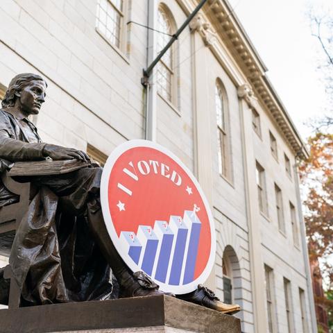 The John Harvard statue with a giant "I Voted" foam core medallion propped up against the statue's legs in Harvard Yard.