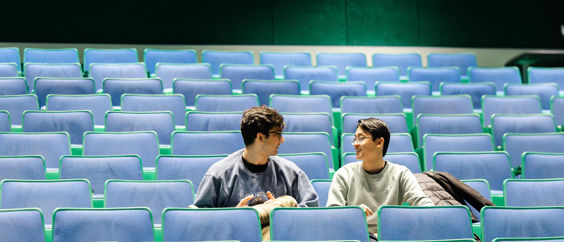 Two students sit in empty lecture hall seating as they talk to each other.