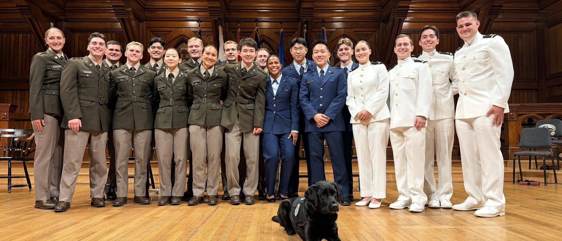 The 2026 ROTC Joint Commissioning candidates stand on stage after receiving their commissions in Sanders Theatre with Sasha the community engagement dog.