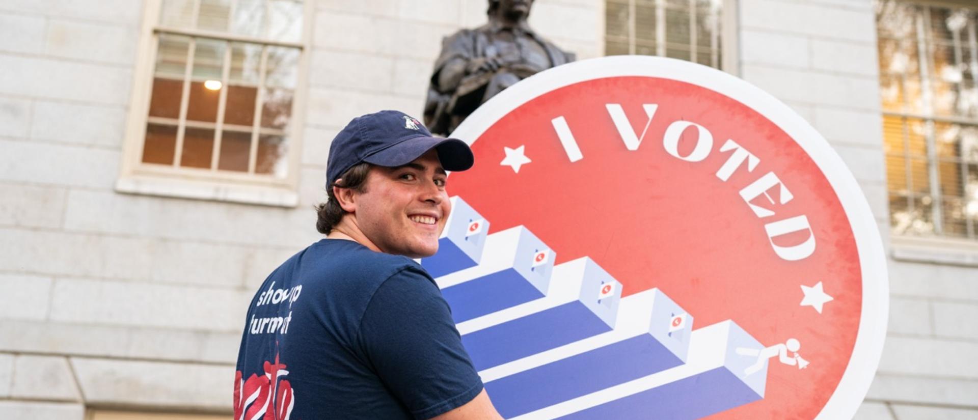A student holds a giant circular "I Voted" sign in front of the John Harvard statue smiling as they look back at the camera.