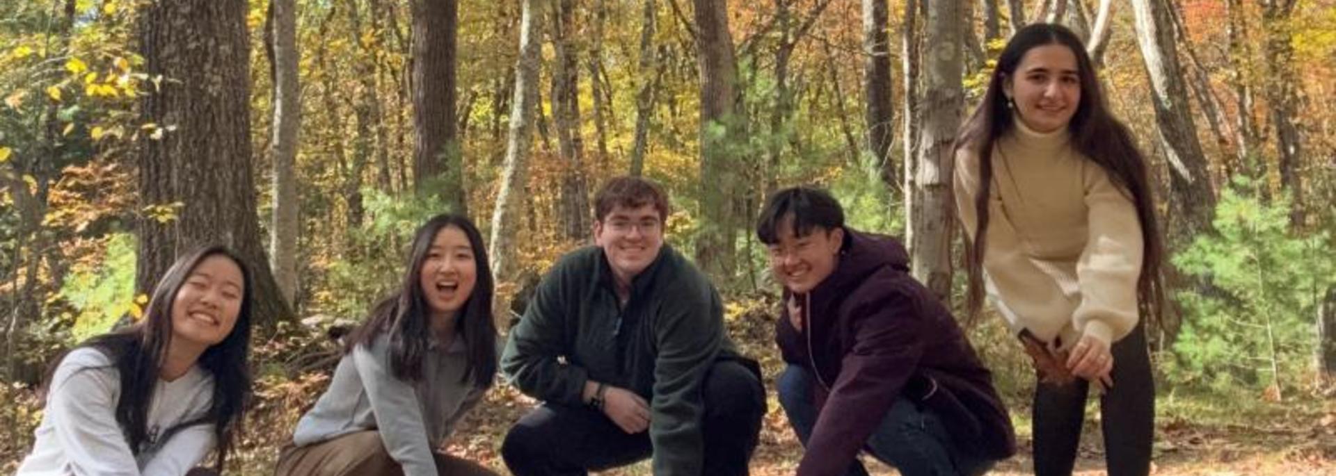 Five students crouch down near the ground for a photo in the Harvard Forest during a sustainability retreat.