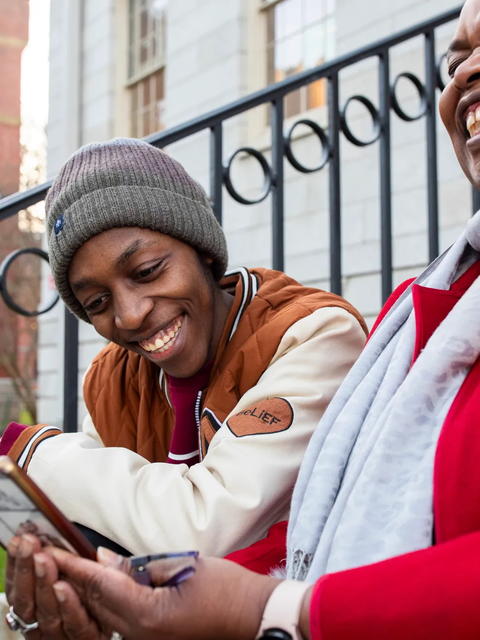 A student sits on University Hall steps with their host parent, Dean Sheila Thimba, as they laugh over a book.