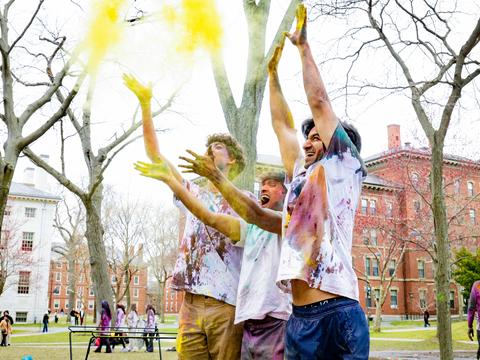 Three students lined up next to each other and throw yellow powders up in the air during the 2025 Holi celebration.