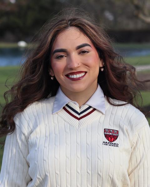 Allyssa Matias, smiling wearing a Harvard Law School sweater, stands outdoors in front of a pond and trees on campus