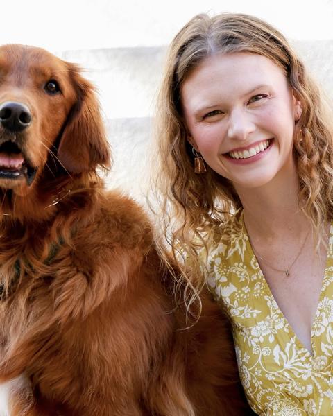 "Jess Cmiel, smiling in a yellow floral dress, sits beside a golden retriever on outdoor steps."