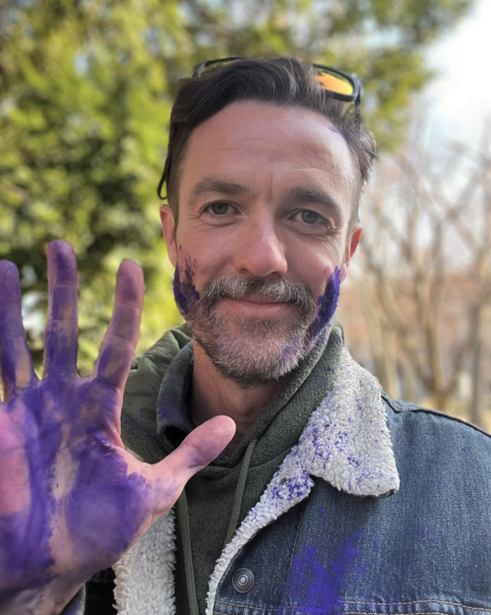 Michael Ferguson smiles at the camera with purple paint on his hand and face, outdoors on a sunny day