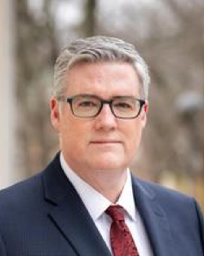 Man with gray hair and glasses in a suit standing outside looking at the camera.